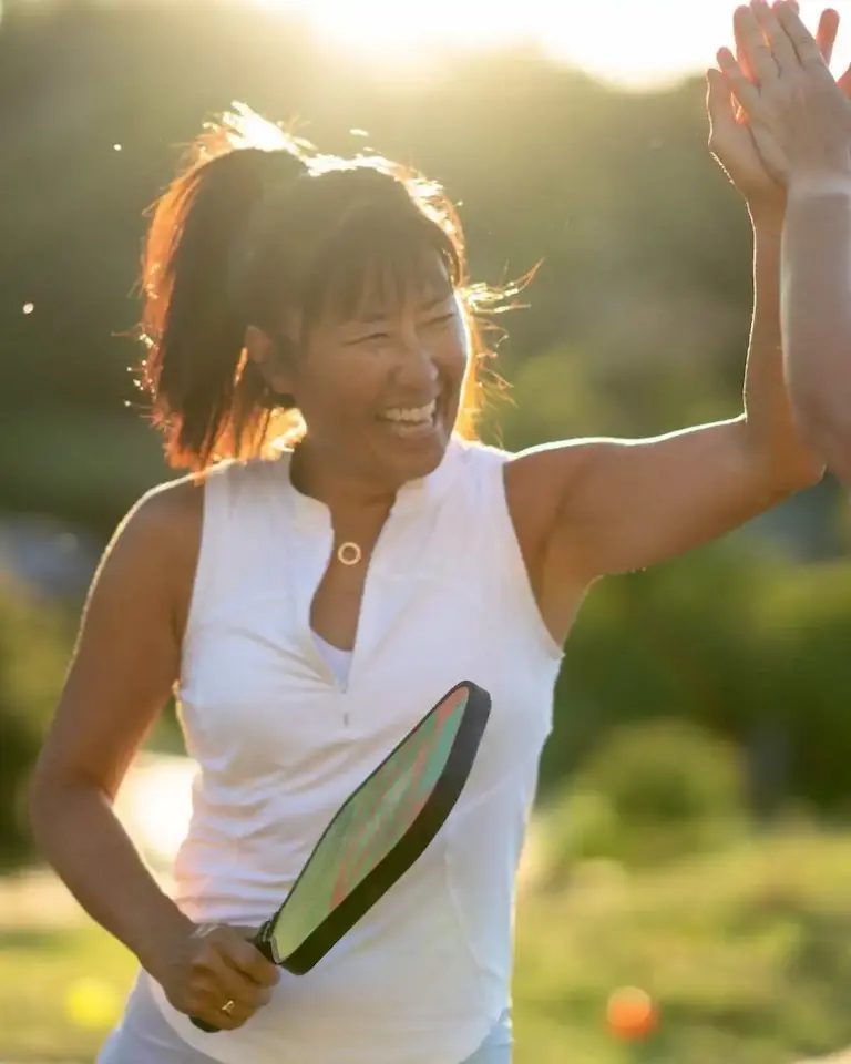 Personne souriante jouant au pickleball en extérieur, raquette à la main, donnant un high five à sa partenaire
